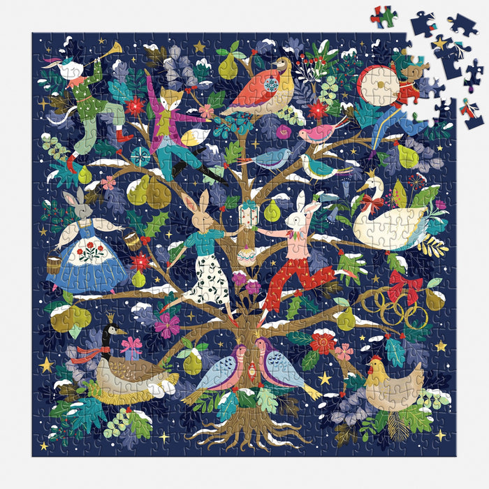 The Flora Waycott "12 Days of Joy" 500 Piece Foil Puzzle features animals and people in costume dancing in a tree on a dark blue background, with some missing pieces at the top right revealing the puzzle’s incomplete state.