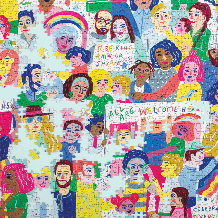The All Are Welcome Here! 1000 Piece Family Puzzle by Sarah Walsh features illustrated people with BE KIND RAIN OR SHINE and ALL ARE WELCOME HERE signs. Some pieces are missing, creating small gaps in the uplifting scene.