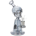 A clear glass skull water pipe with a metallic silver base and a glass bowl attachment, featuring a black logo on the neck, set against a plain white background.