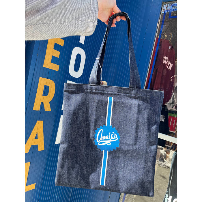 Denim tote bag with a brand logo held by a person against a store entrance.