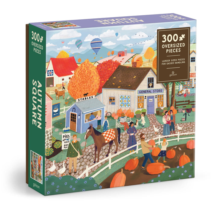 The Elisabeth Dorrian Autumn Square 300 Piece Puzzle features an autumn village scene with people, horses, pumpkins, colorful trees, a general store, stables, and hot air balloons. The box reads “Autumn Square.”.