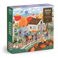 The Elisabeth Dorrian Autumn Square 300 Piece Puzzle features an autumn village scene with people, horses, pumpkins, colorful trees, a general store, stables, and hot air balloons. The box reads “Autumn Square.”.
