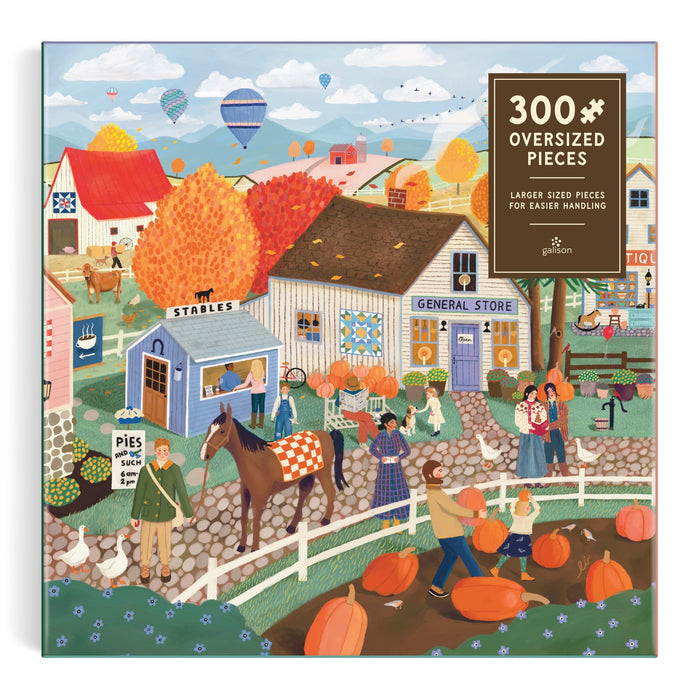 The Autumn Square 300 Piece Puzzle by Elisabeth Dorrian features a vibrant fall village with pumpkin pickers, a horse by stables, hot air balloons above, and colorful trees. The box states “300+ oversized pieces.”.