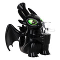 A black dragon figurine with glowing green eyes holding a clear glass water pipe, set against a plain white background.