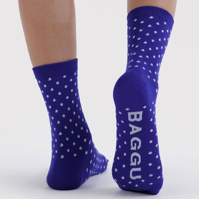 A pair of royal blue crew socks with white polka dots and the brand name 'BAGGU' printed vertically in white on the sole, shown on a person's feet against a light gray background.