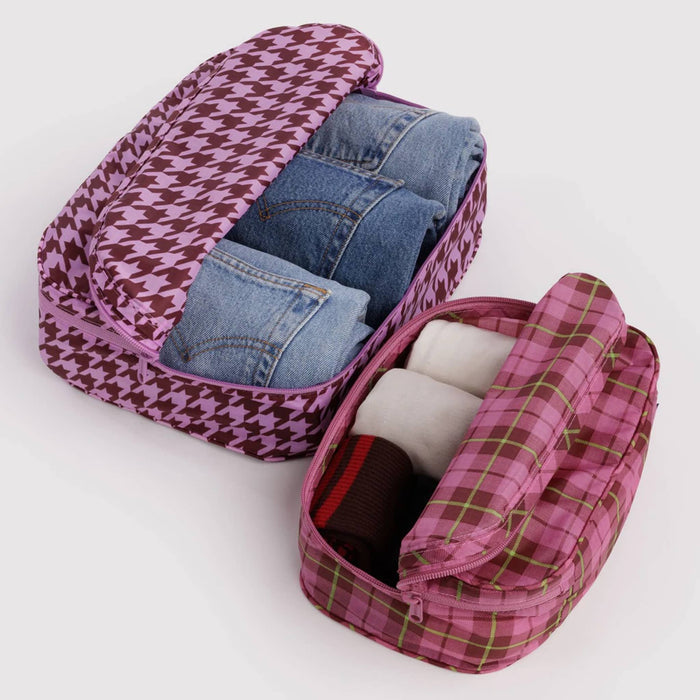 2 recycled nylon zippered packing cubes, large bag is pink and brown houndstooth with jeans inside, smaller bag is pink tartan with socks inside, on a white background.