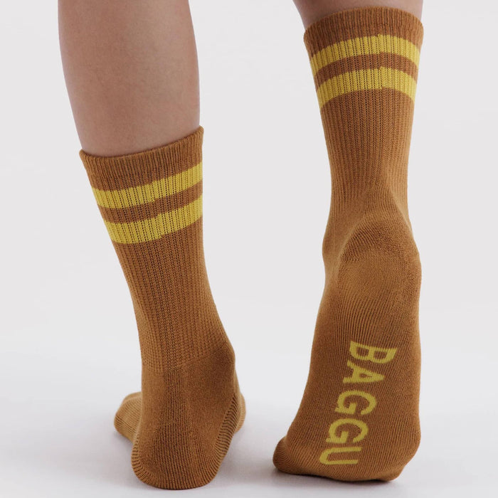 A pair of brown crew socks with yellow stripes near the top and the brand name 'BAGGU' printed in yellow on the sole, shown worn on a person's feet against a light gray background.