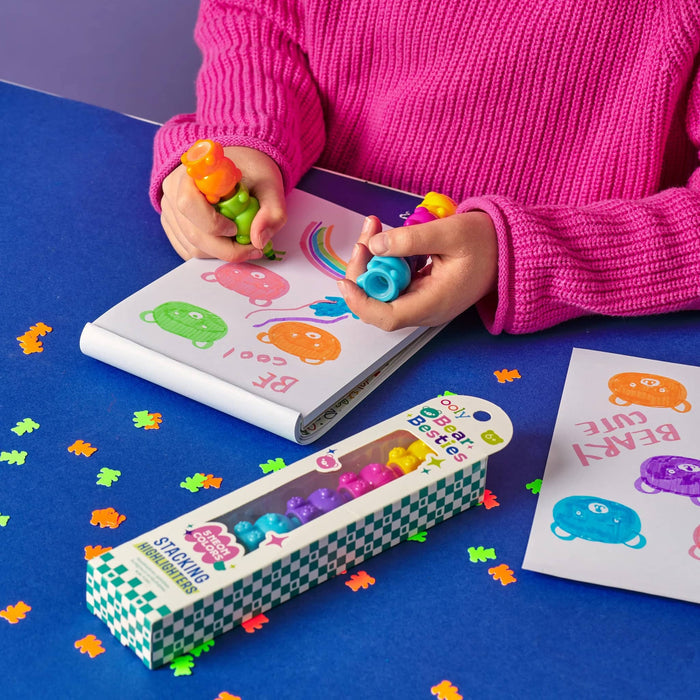 A child wearing a bright pink sweater drawing with bear-shaped highlighter markers in a white notebook on a blue table. A boxed set of highlighters and bear shaped confetti are also visible on the table.