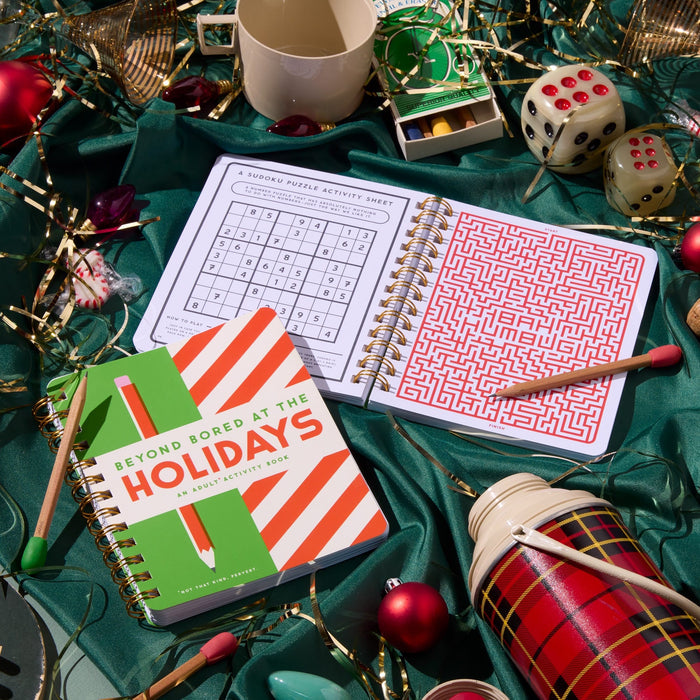 A festive flat lay displays open Beyond Bored At The Holidays puzzle books by Brass Monkey, a striped journal, ornaments, dice, a thermos, string lights, and decorations on green fabric—ideal for lovers of holiday brain games!.