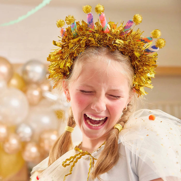 Young girl wearing a gold tinsel headband with colorful fabric candles and pom-poms in a festive setting with balloons.