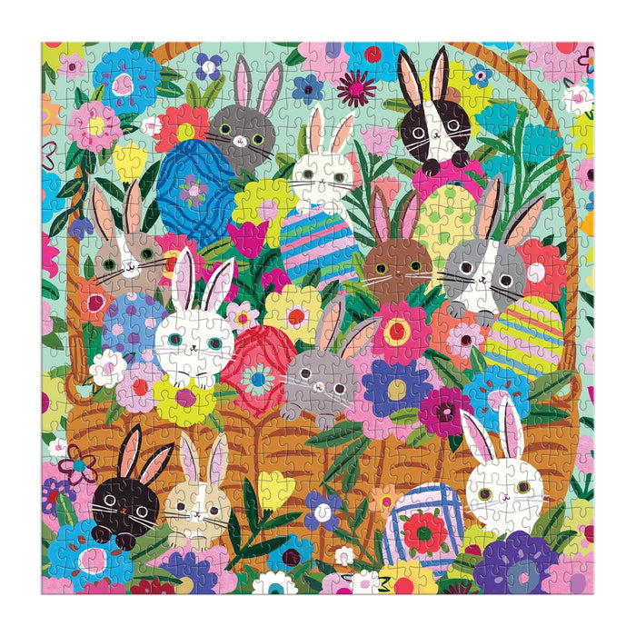 The Blooming Bunnies 500 Piece Foil Family Puzzle by Tara Lilly features a vibrant illustration of adorable bunnies, a basket of Easter eggs, and colorful flowers and green leaves in eye-catching detail.