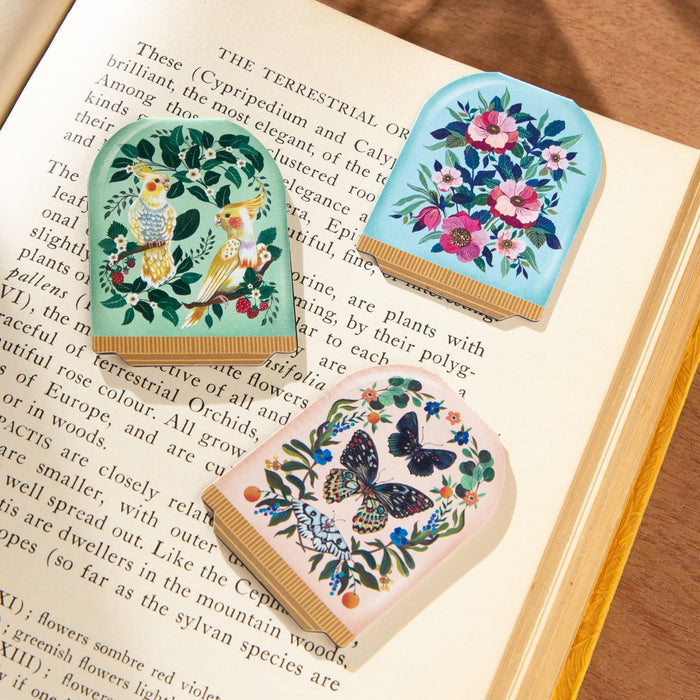 Three Blooming Terrariums Magnetic Bookmarks by Christine De Carvalho—featuring owls, flowers, and a butterfly—adorn the open pages of The Terrestrial Orchids on a wooden surface.