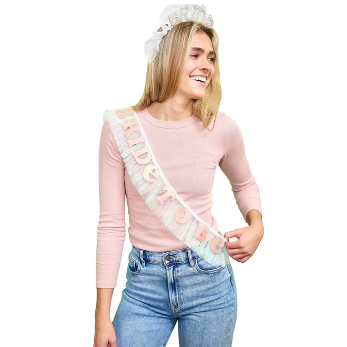 Woman wearing an iridescent white fringe sash with the words 'BRIDE TO BE' in shiny rose gold letters along with a headband against a plain white background.