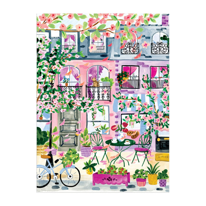 The Cherry Lane Blossoms 1000 Piece Puzzle by Sabina Fenn features a vibrant pink and purple building with balconies, blooming trees, café seating, a bicycle, and lush plants, capturing a cheerful street scene.
