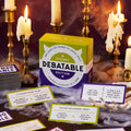 A tabletop display features Brass Monkey's Debatable Spooky Edition cards and game box, surrounded by flickering candles, fake spiderwebs, and quirky debate questions in a dark, atmospheric setting.
