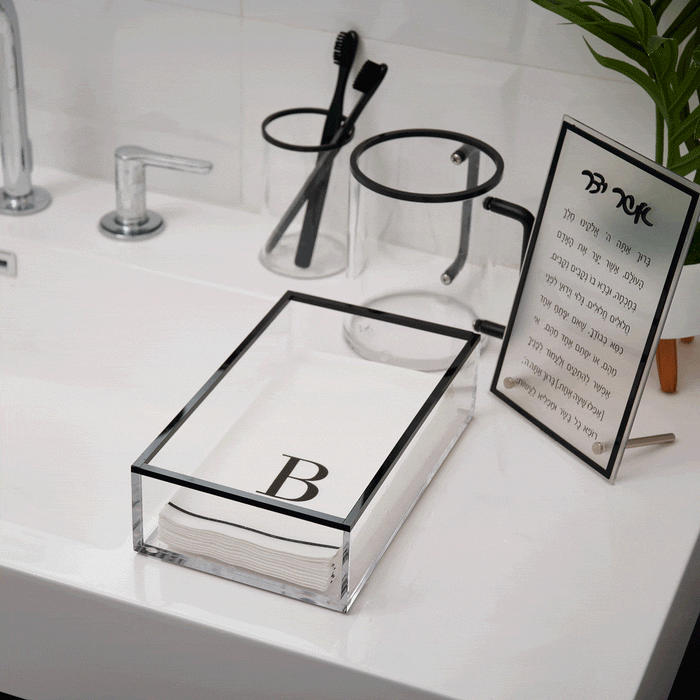 Clear acrylic guest towel holder with black trim frame displaying the letter B, styled on a white bathroom vanity with toiletries and decor.