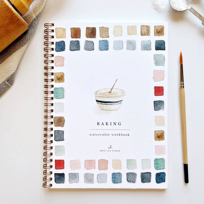 Front cover of the Baking Watercolor Workbook on a white surface with a paintbrush and measuring spoon.