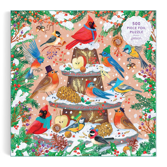 The Festive Feathers 500 Piece Foil Puzzle by Olga Akbarova showcases cardinals and robins on a snowy bird feeder, surrounded by pine branches, berries, pinecones, and apples on a pink backdrop.