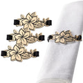 Four gold floral napkin wraps with black bands arranged on white napkin, showing detailed flower designs from Waterdale Collection.