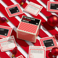 A festive flat lay displays Brass Monkey's Frivolous Holiday Trivia game box, trivia cards, and red ornaments on a striped background—perfect for holiday parties.