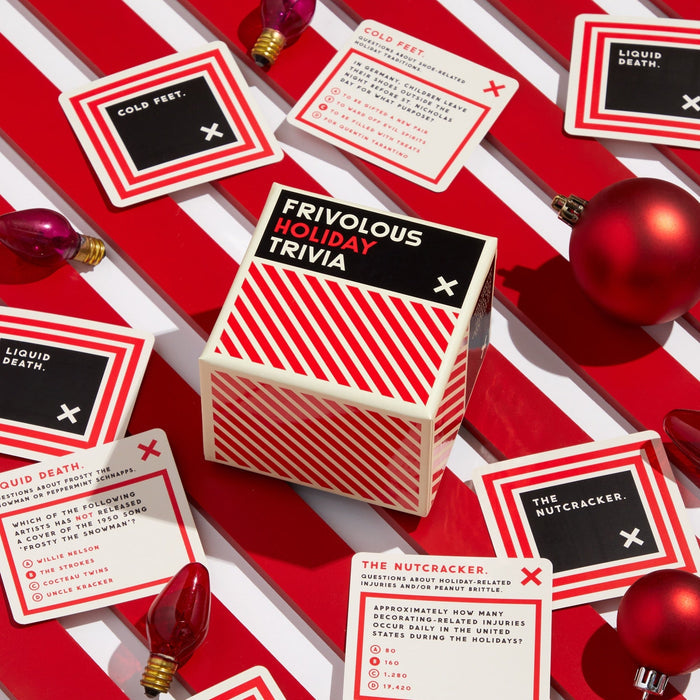 A festive flat lay displays Brass Monkey's Frivolous Holiday Trivia game box, trivia cards, and red ornaments on a striped background—perfect for holiday parties.
