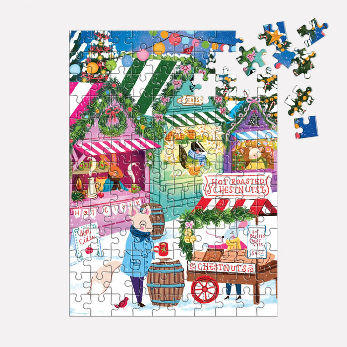 The Joanna Clay Holiday Market 130 Piece Puzzle Ornament shows a festive outdoor holiday market scene with decorated stalls, hot cocoa and roasted chestnut signs, shoppers, and several loose pieces visible in the top right corner.