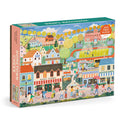 Hometown Cheer 1000 Piece Puzzle by Elisabeth Dorrian features a vibrant small town scene with shops, a pizzeria, market stalls, people strolling, trees, and houses.