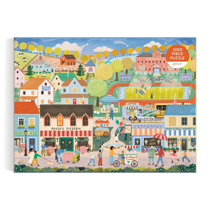 Box art for the Hometown Cheer 1000 Piece Puzzle by Elisabeth Dorrian, featuring a vibrant small town with colorful buildings, shops, people strolling, a pizzeria, market, park, and a distant football field.