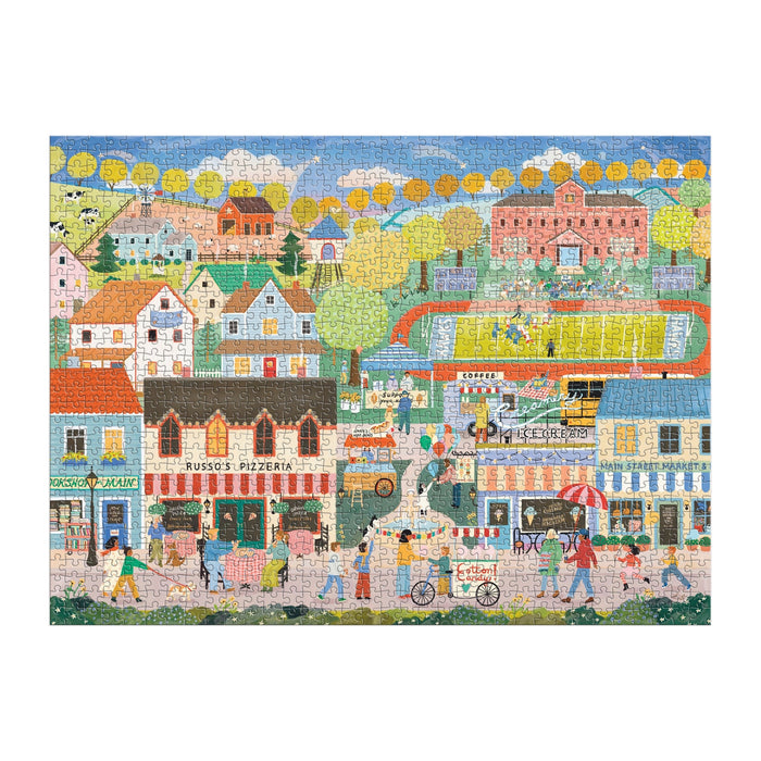 Hometown Cheer 1000 Piece Puzzle by Elisabeth Dorrian features a vibrant small town scene with shops, a pizzeria, homes, people walking, cycling, and playing soccer amid trees and a large building.