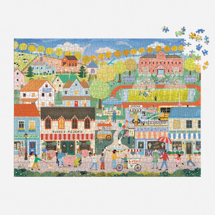 The Elisabeth Dorrian Hometown Cheer 1000 Piece Puzzle features a vibrant town scene with shops, houses, people outdoors, bikers, and a soccer game in the stadium. Some pieces remain to be placed for the full picture.
