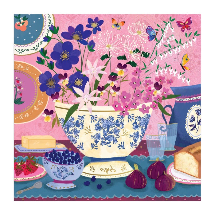 The Joy Laforme Art of the Table 500 Foil Piece Puzzle features a vibrant still life with flowers in a blue and white vase, fruit, bread, butter, and decorative plates on a pink tablecloth. Brand: Joy Laforme.