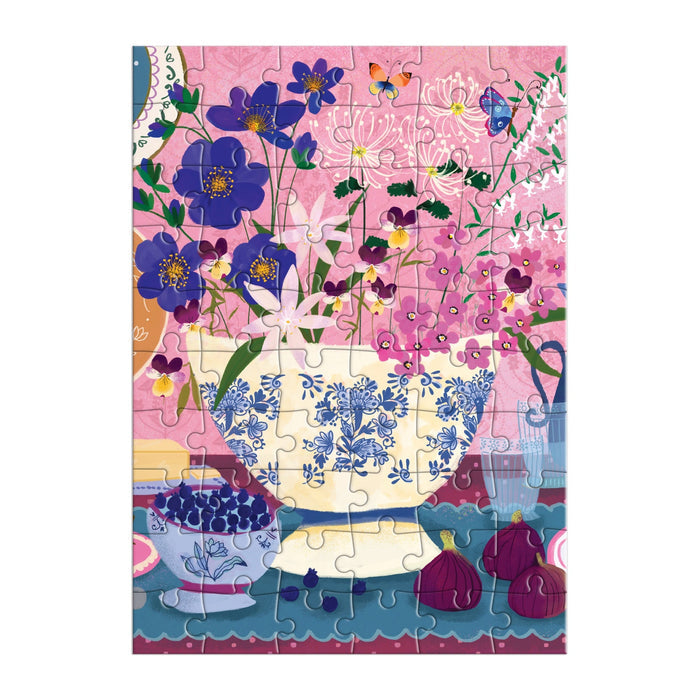 The Joy Laforme Art of the Table Greeting Card Puzzle by Joy Laforme features a blue-and-white floral vase with colorful flowers, a bowl of blueberries, figs on a table, and a pink patterned background with butterflies.