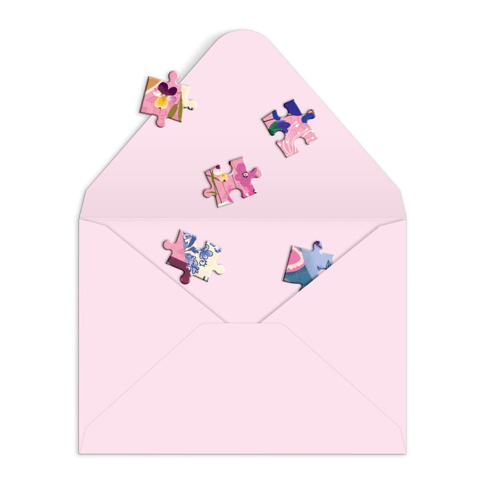 Colorful jigsaw pieces spill from a pink envelope on a white background in the Joy Laforme Art of the Table Greeting Card Puzzle by Joy Laforme.