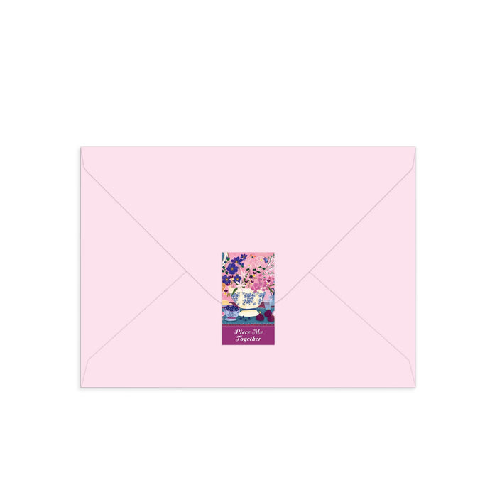 The Joy Laforme Art of the Table Greeting Card Puzzle by Joy Laforme comes in a light pink envelope with a back flap sticker, adorned with pink and blue floral art and “Peace, Not Together” at the bottom.