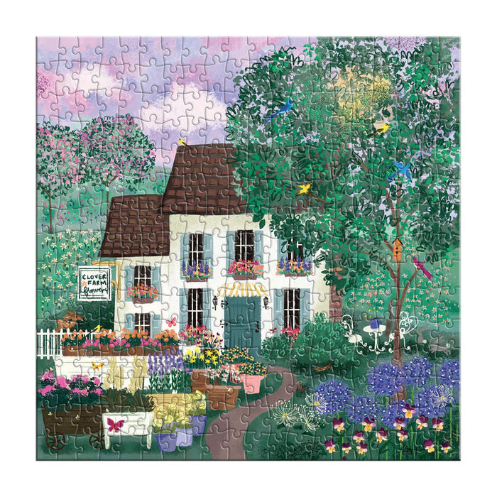 The Joy Laforme Garden Path 300 Piece Puzzle by Joy Laforme features a white house with a brown roof, colorful flowers, green trees, flowering bushes, birds, and a Clover Farm sign under a purple-blue sky.