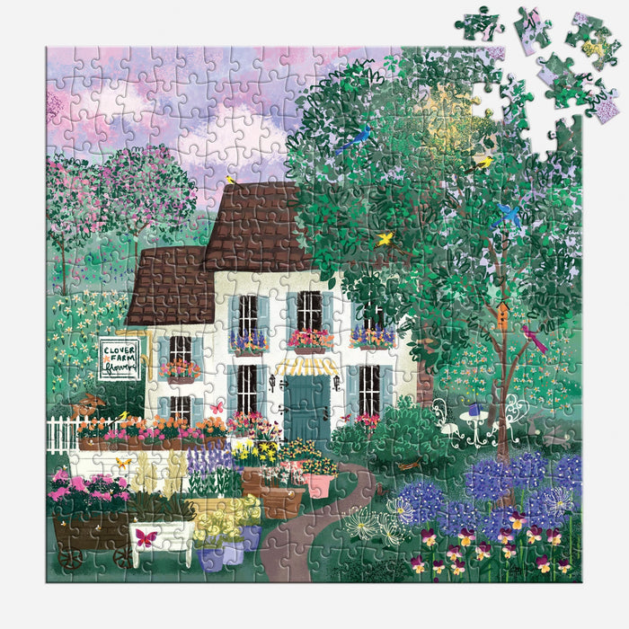The Joy Laforme Garden Path 300 Piece Puzzle by Joy Laforme features a nearly finished scene of a white cottage with a brown roof amid vibrant flowers and green trees, under a pink and purple sky. A few pieces are missing from the top right corner.