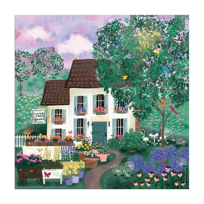 The Joy Laforme Garden Path 300 Piece Puzzle by Joy Laforme features a charming house with a brown roof, vibrant flowers, lush trees, butterflies, flower carts, birds, and a sign reading Clover Flower Garden.