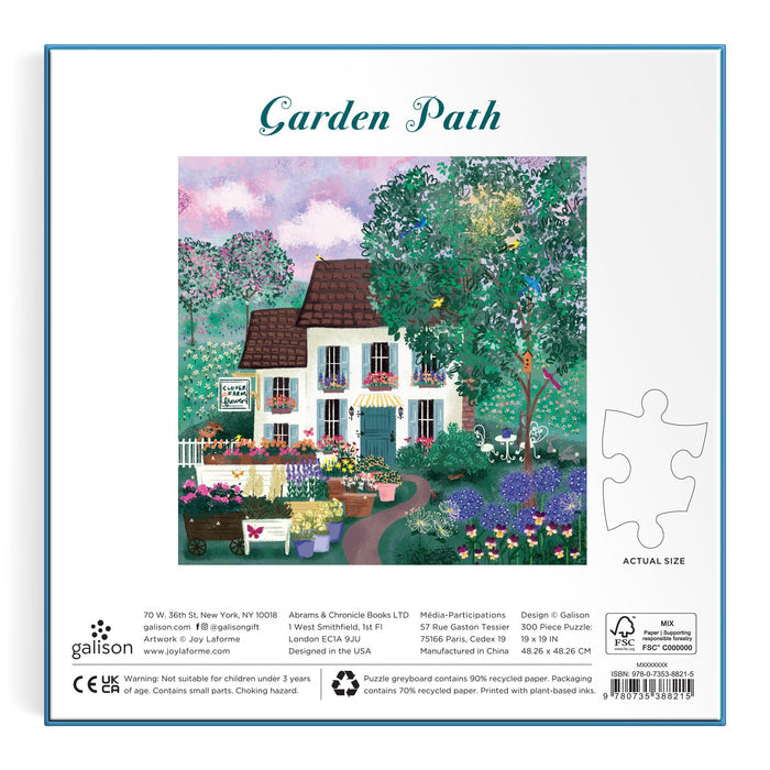 The image shows the back of the Joy Laforme Garden Path 300 Piece Puzzle box, featuring artwork of a charming white house, colorful flowers, trees, a fountain, and garden path. "Garden Path" appears above the scene. Brand: Joy Laforme.