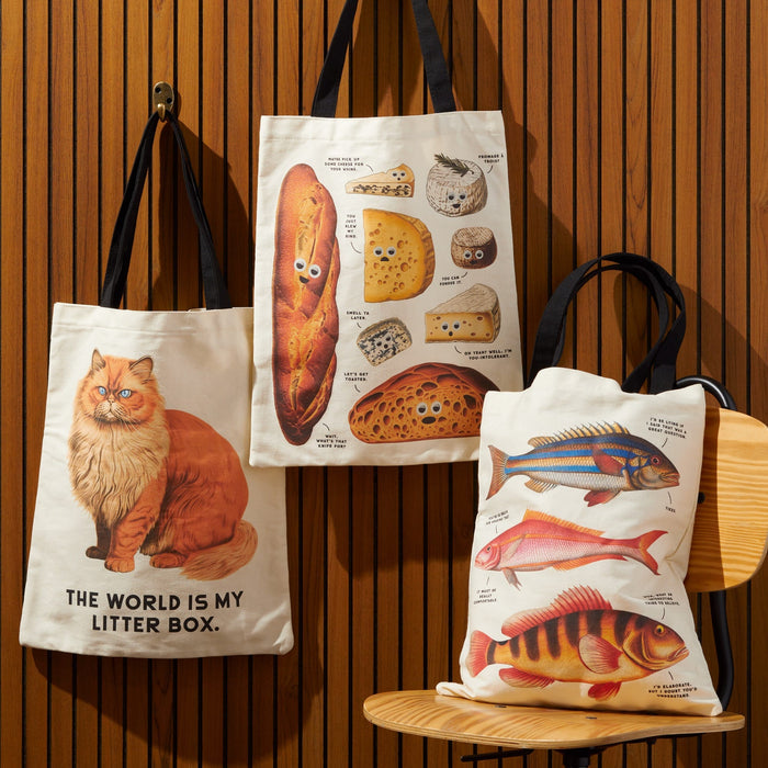 Three reusable Brass Monkey tote bags hang on a wooden wall and chair: one with an orange cat and funny saying, another labeled breads, and the Judgy Fish Canvas Tote featuring colorful vintage fish artwork.