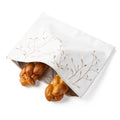 White linen challah cover with delicate tan leaf embroidery, showing roasted challah bread spilling out beside it on white background.