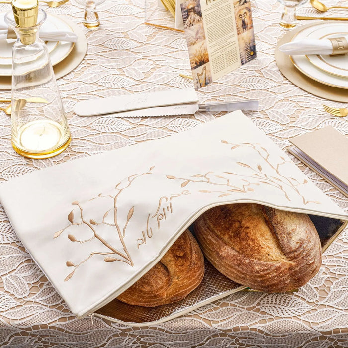 White challah cover with delicate gold leaf embroidery, displayed on a table setting with bread and place settings for Shabbat dinner.
