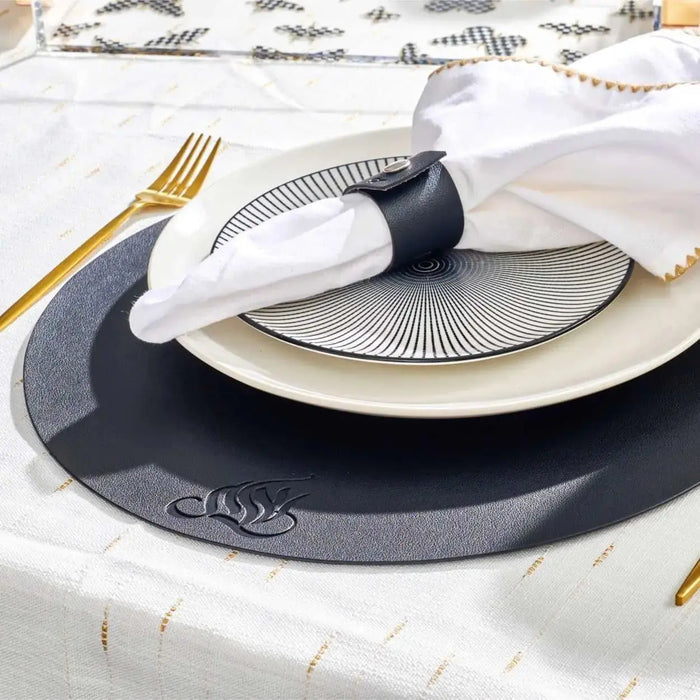 Black leather round charger plate with silver border detail, styled with white napkin, gold fork, and place setting on white tablecloth.