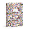 The Liberty London Nell, Annie and May Premium A5 Notebook is a hardcover A5 notebook with a vibrant red, blue, yellow, and green floral pattern and a white label reading “LIBERTY. A5 NOTEBOOK 300 PAGES.”.