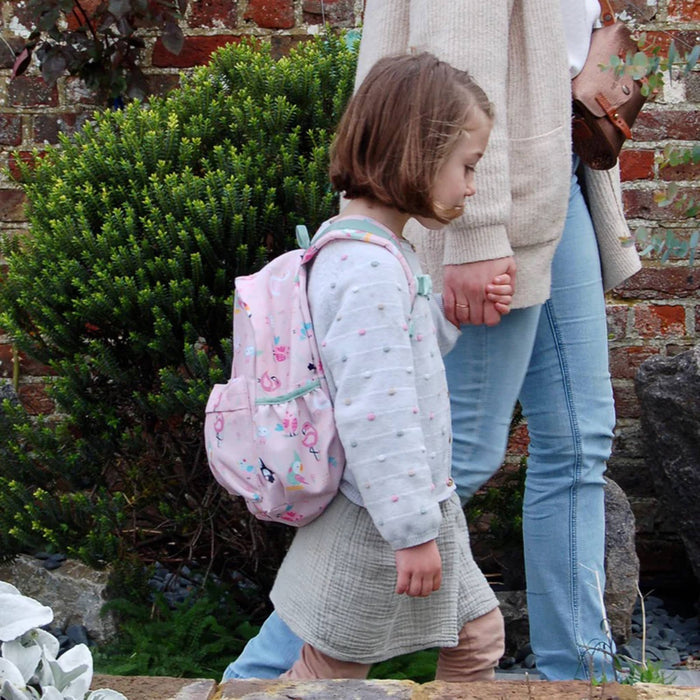A young girl with short brown hair walks outdoors wearing a light pink patterned backpack, a light gray long-sleeve top, and a gray skirt, while holding hands with an adult wearing a beige sweater and blue jeans.