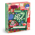 The Mahjong Social 1000 Piece Puzzle by Paige Jung showcases a vibrant illustration of a Mahjong game table with tiles, tea, snacks, flowers, and cozy decor in pink, green, and red hues.