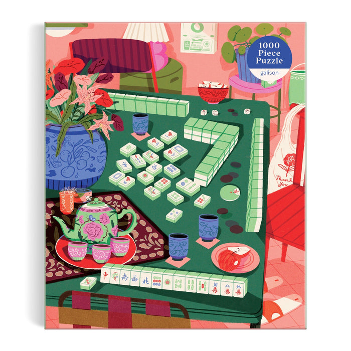 The Mahjong Social 1000 Piece Puzzle by Paige Jung showcases a vibrant mahjong game on a green table, complete with a floral tea set, vases of flowers, and decorations in a cozy, warmly colored room.