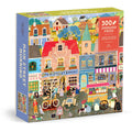 The Elisabeth Dorrian Main Street Morning 300 Piece Puzzle features oversized pieces and a vibrant street scene with shops, a bakery, café, flowers, and people enjoying the day.