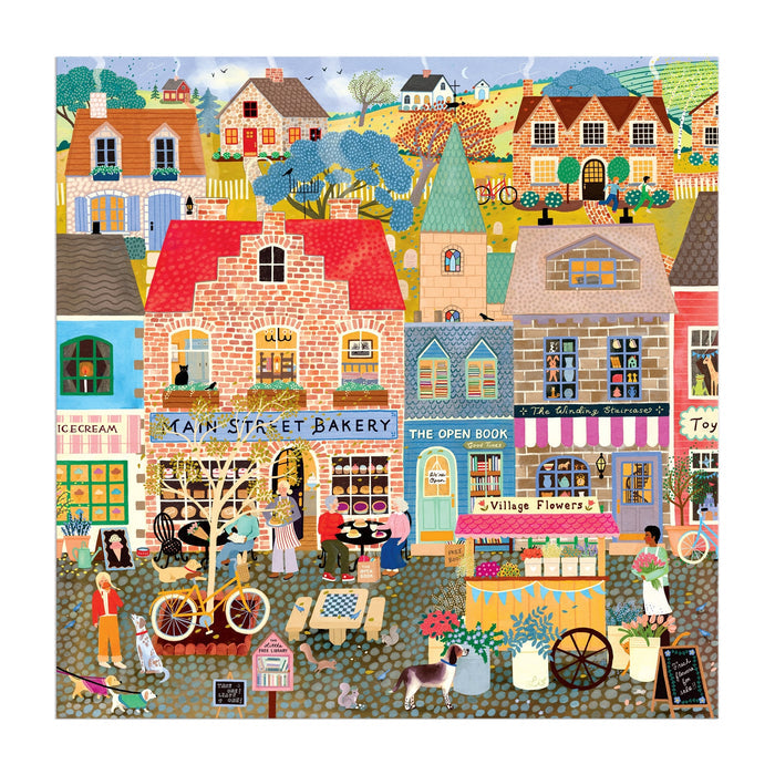 Main Street Morning 300 Piece Puzzle by Elisabeth Dorrian features a vibrant village with shops—a bakery, bookstore, ice cream shop, and flower stand—plus people walking, cycling, chatting, kids playing, a dog, and bright houses with hills in the background.