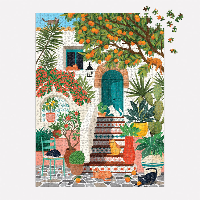 The Mediterranean Cats 1000 Piece Puzzle by Tània Garcia features a vibrant courtyard scene with lush plants, a tiled staircase, cats relaxing among pots and steps, and an overhanging orange tree. A few pieces are unfinished in the upper right corner.