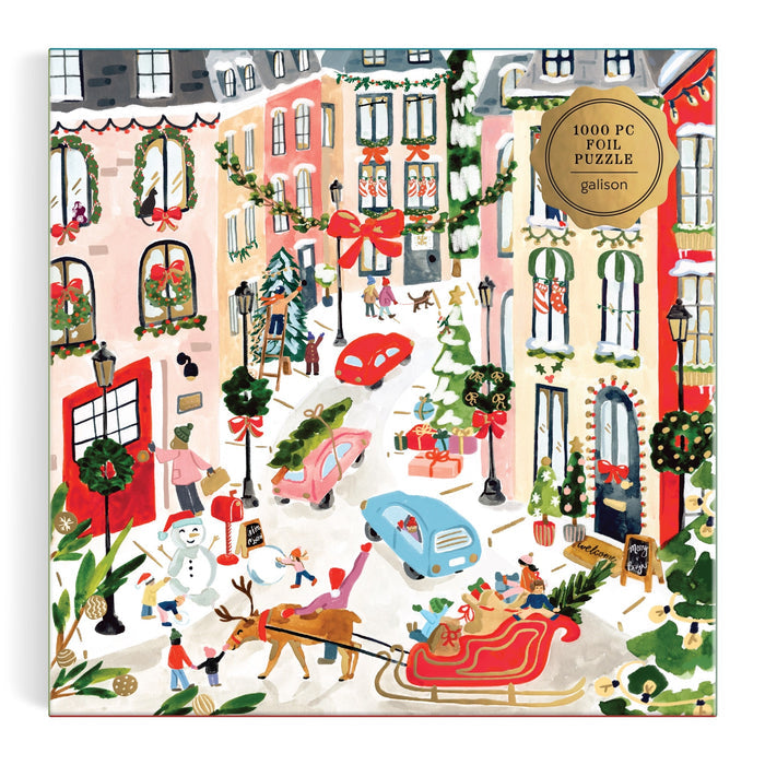 Festive city street puzzle features decorated buildings, people, a dog-pulled sleigh, gifts, Christmas trees, and cars; gold label in the corner reads “Merriest Village 1000 Piece Foil Puzzle Sabina Fenn.”.